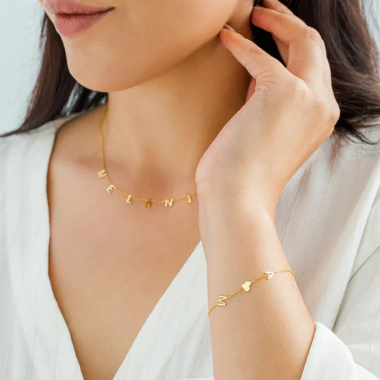 Een elegante close-up van een vrouw die een gouden Double Love Heart Bracelet draagt, gecombineerd met een bijpassende ketting met letters. De subtiele armband heeft twee hartvormige details, perfect voor een tijdloze en stijlvolle look. Ideaal om elegantie en een symbolische touch aan elke outfit toe te voegen.